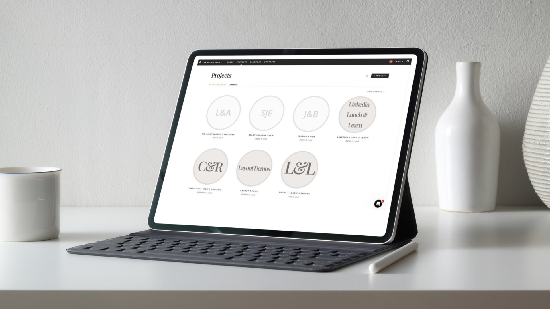 Project Image A professional tablet with a keyboard displays the Aisle Planner Pro wedding planning software and portfolio on a white desk, alongside a coffee mug and digital stylus for a modern event coordinator workspace.